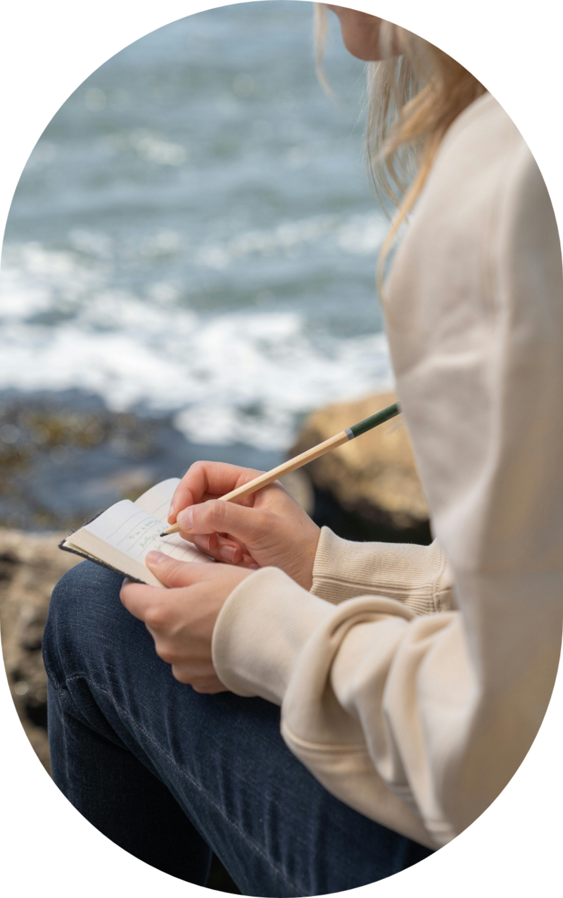 Woman sitting by the ocean writing in a notebook, representing reflection, self-discovery, and the therapeutic process of reconnecting with oneself through feminist, values-based therapy.