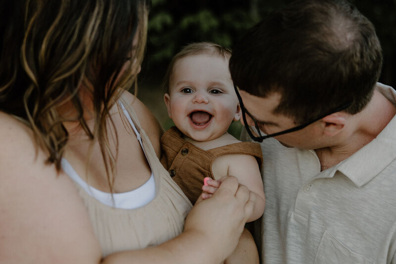 Adorable family photos taken outdoors on a beach with forest in the background, mom is holding baby girl and looking at her, dad is next to them looking at baby girl too who is smiling wide
