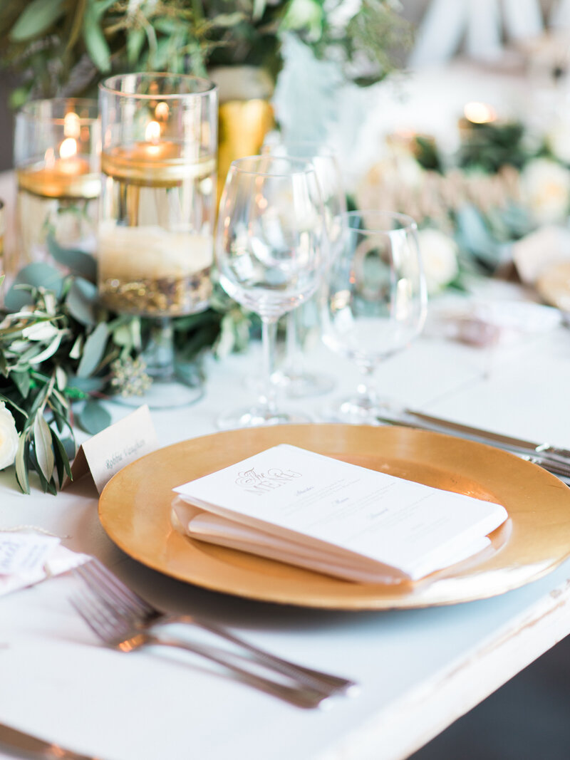 Wedding dinner place setting with serving and flatware, glasses, greenery, and candles