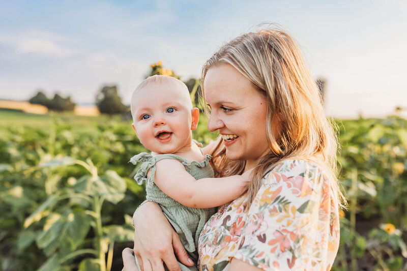 Mom holding her baby girl smiling in a beautiful glowing sun drenched sunflower field for her harrisburg family photo session