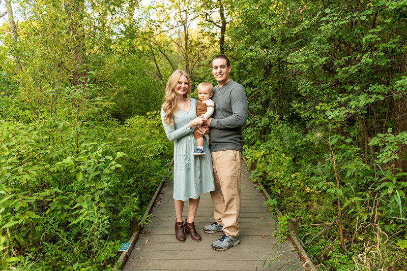 A mom smiles big while holding her toddler son on her hip with dad hugging on a boardwalk in a park