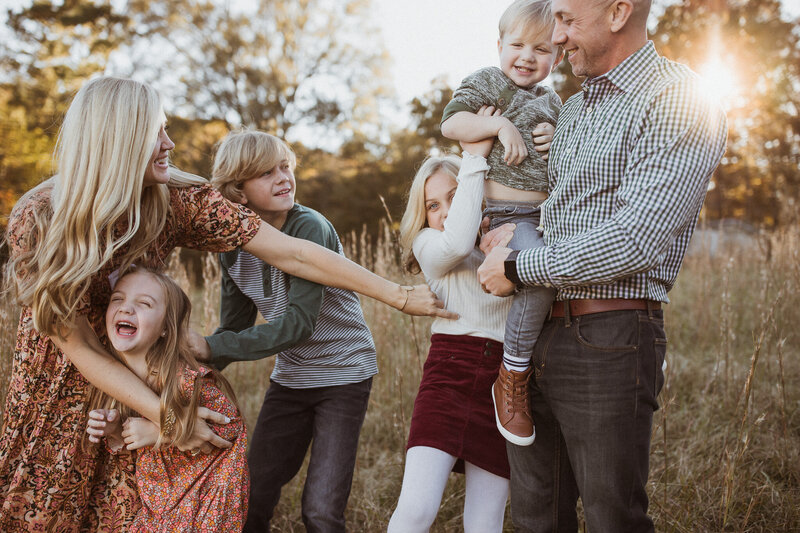 A mom wearing a pretty fall dress tickles her four children while the dad laughs. 