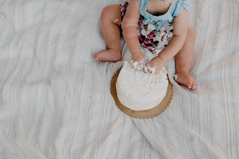 Milestone picture of baby girl digging her hands into cake on white blanket.