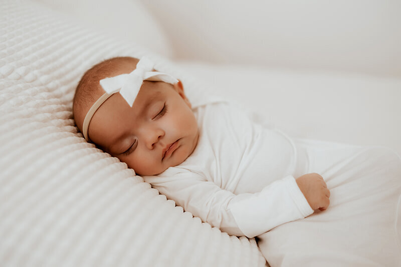 Newborn baby girl in a white sleeper and white bow lays on a white blanket for a Denver newborn session