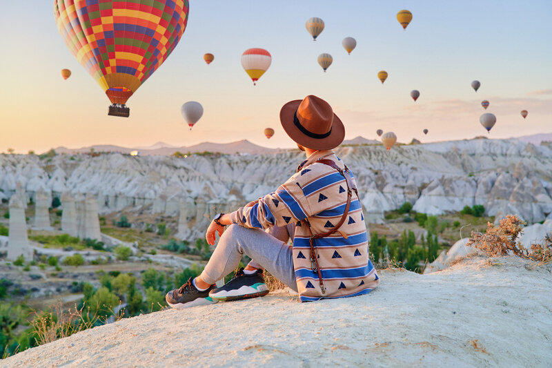 A traveler sits on a rocky ledge, wearing a hat and casual clothing, gazing at colorful hot air balloons floating above a stunning landscape at sunset.