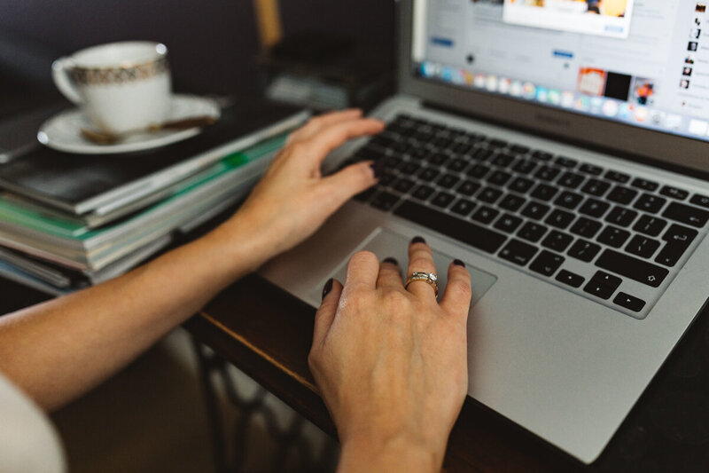 Surrounded by files and papers, a woman works on her laptop. Her coffee sits forgotten beside her.