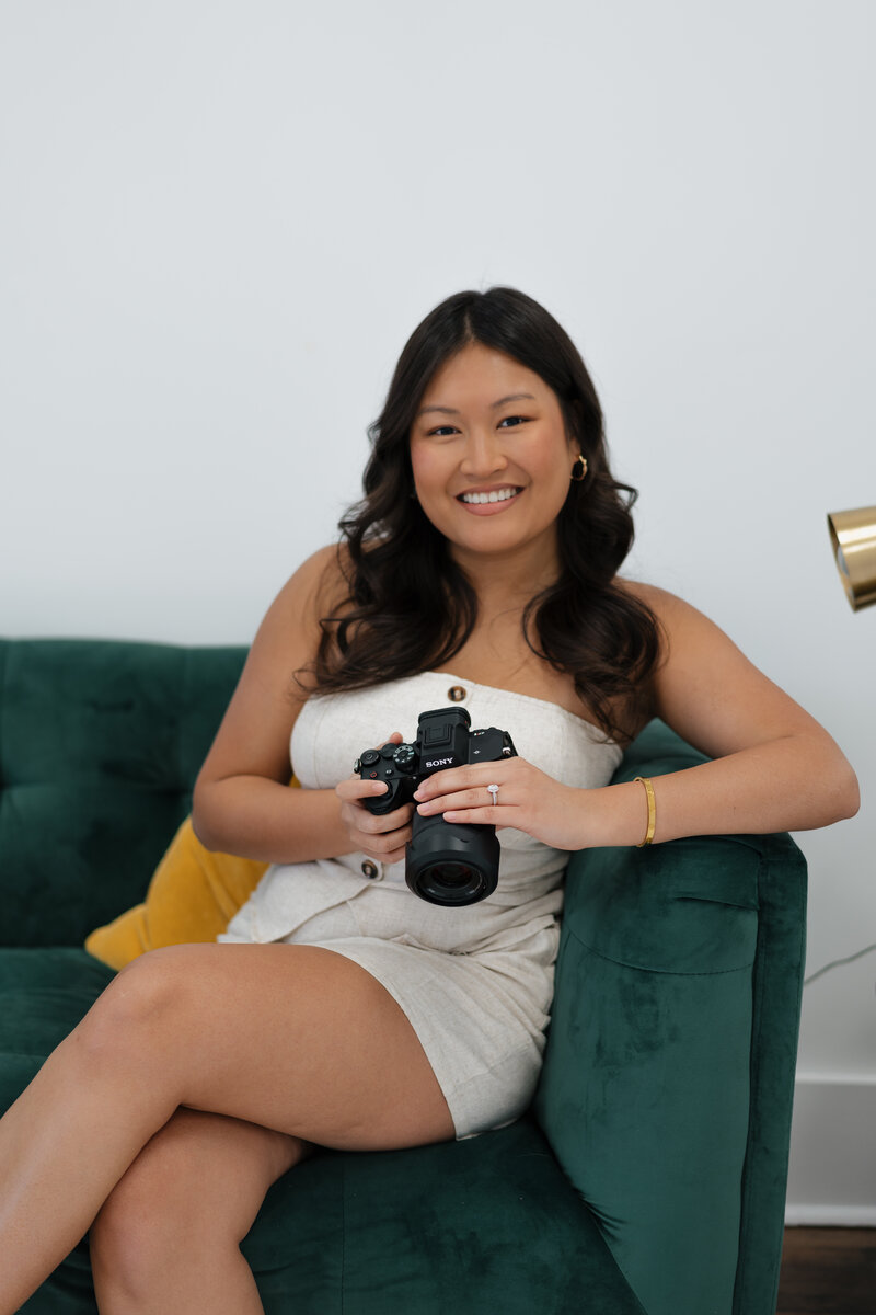 Owner and lead photographer of Pictures by Pim. Pim is holding a camera while sitting and posing on a green couch.