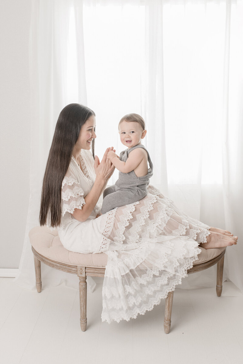 Woman in white lace dress holding hands with one year old baby in front of window