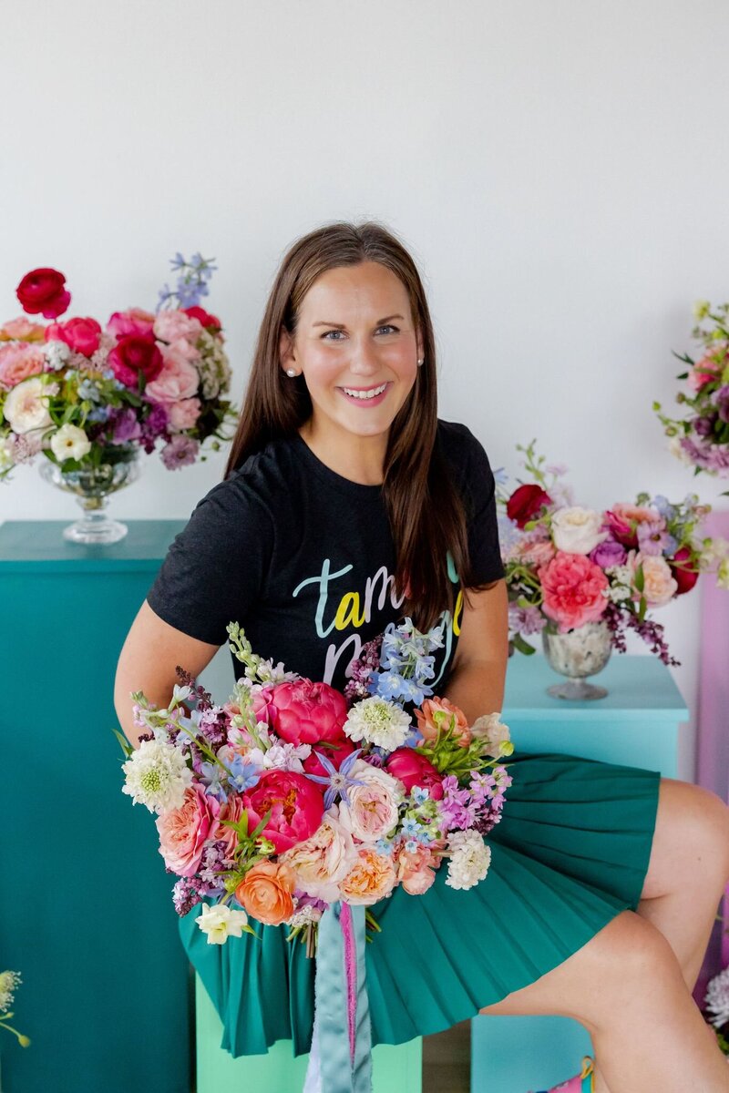 Tamara Menges holding a vibrant garden-style bridal bouquet with colorful blooms, smiling in studio with floral arrangements.