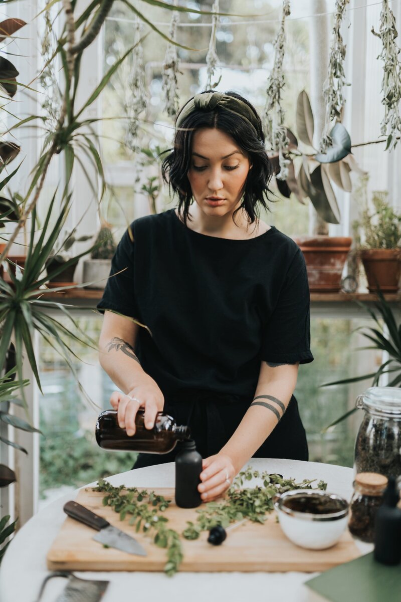 Woman wearing black shirt pours liquid from large amber bottle into small black bottle with a sticker that reads "Moss Herbalism" with a fern illustration above it,
