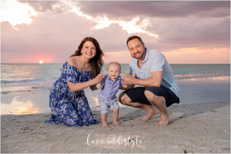 A baby smiles joyfully as his parents hold his hand on the beach at sunset in Sarasota, FL.