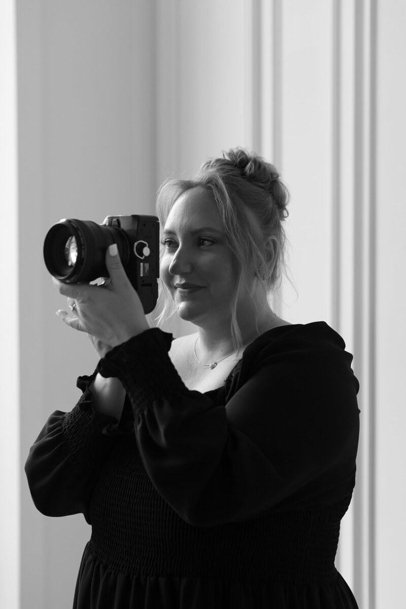 Black and white close up branding portrait of a woman holding a camera and pretending to take a photo