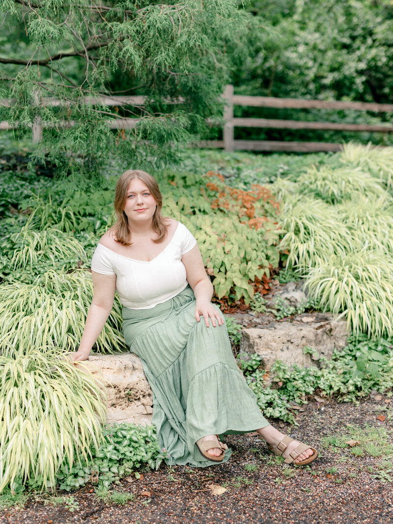 High school senior girl in white top and green maxi skirt sitting amongst nature