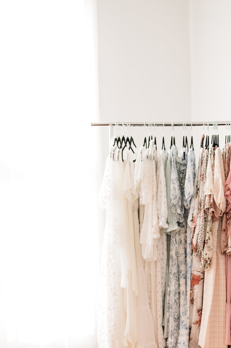 A row of beautiful light colored dresses hanging on a rack in front of a window