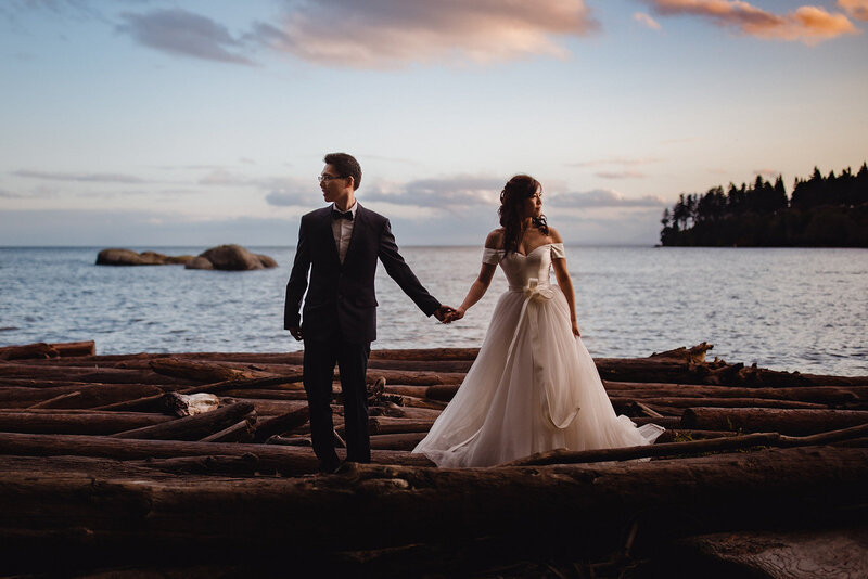 An Asia bride and groom stand hand-in-hand on the beach. The look to opposite sides. The groom wears a black suit, white shirt, and black bow tie. The bride wears a princess-like white ballgown with a sweetheart neckline. They stand on driftwood logs that clutter the shore. Behind them is the ocean with rocks and a small island in the distance. The sky in light blue with a few clouds which are a pink/orange colour.