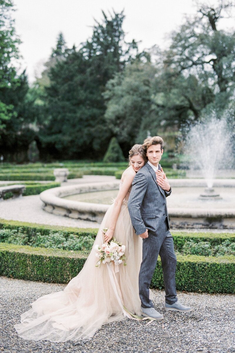 bride-with-bouquet-hugs-groom-from-near-fountain