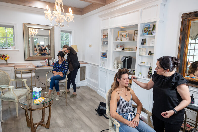 Four women in a room are getting ready for a wedding. One woman is getting her makeup done and one woman is getting her hair done