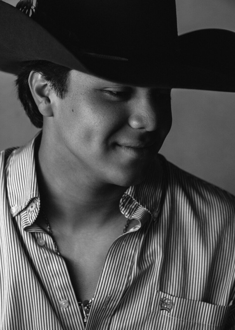 Young man wearing button down shirt and cowboy hat looking down to right with a slight smile on his face. Black and white image of senior Andrew Greene shot in 2024.