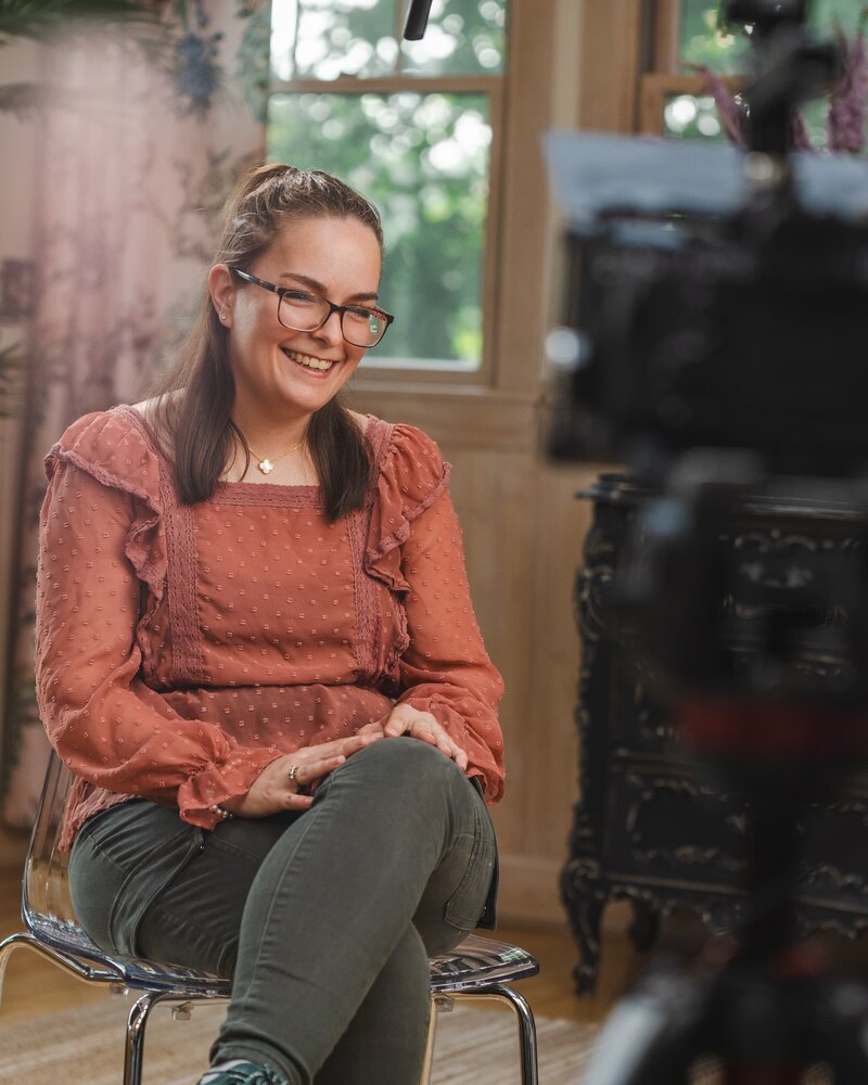 Smiling interview subject with glasses on a clear chair, rust blouse and green pants; boom mic overhead, camera in foreground, window-lit room.