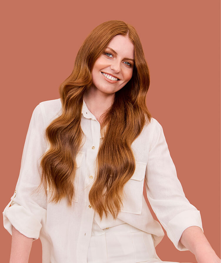 Woman with long, flowing red hair styled in soft waves, wearing a white button-up shirt against a terracotta backdrop.