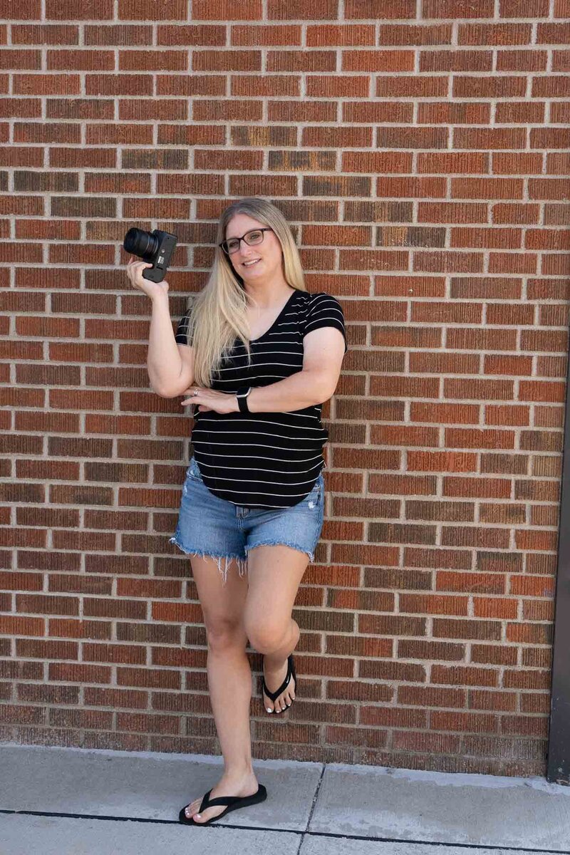 Megan smiling softly while standing against a brick wall holding her camera in both hands | Senior Photographer in Lawrence, KS