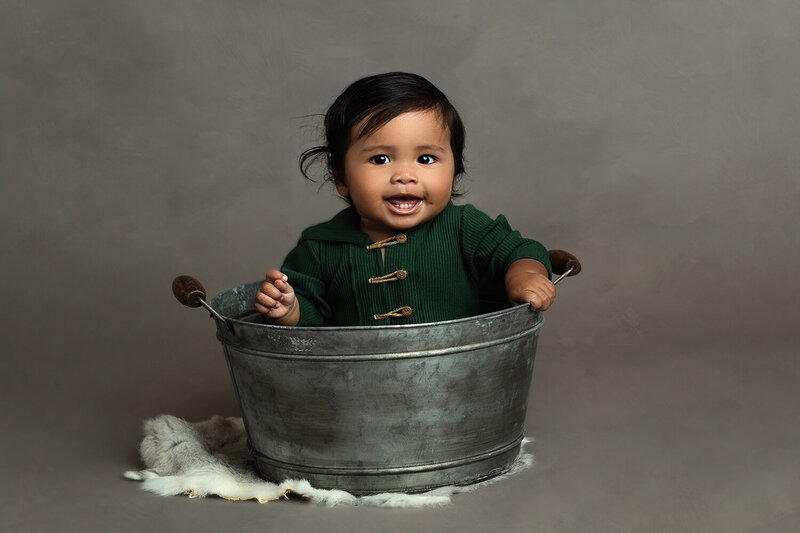 A young toddler sits in a tin bucket in a green sweater in a studio