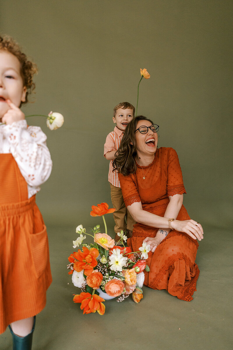 A mother holds flowers and laughs with her children besides her in a photography studio, representing a family photography portraits session. 