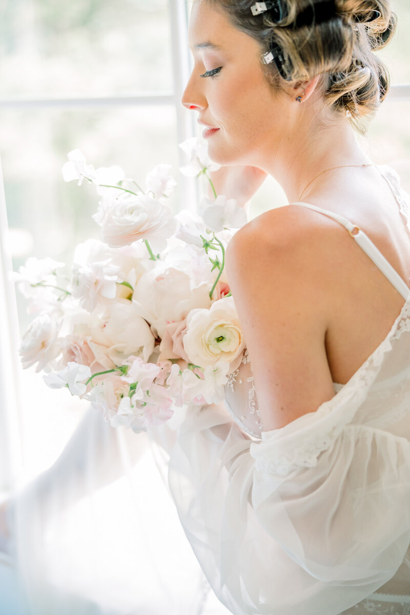 Richmond Boudoir Photography | A closeup of a girl in white floral lingerie and a sheer white robe draped off her shoulders holds a delicate floral bouquet as she sits in a windowsill.