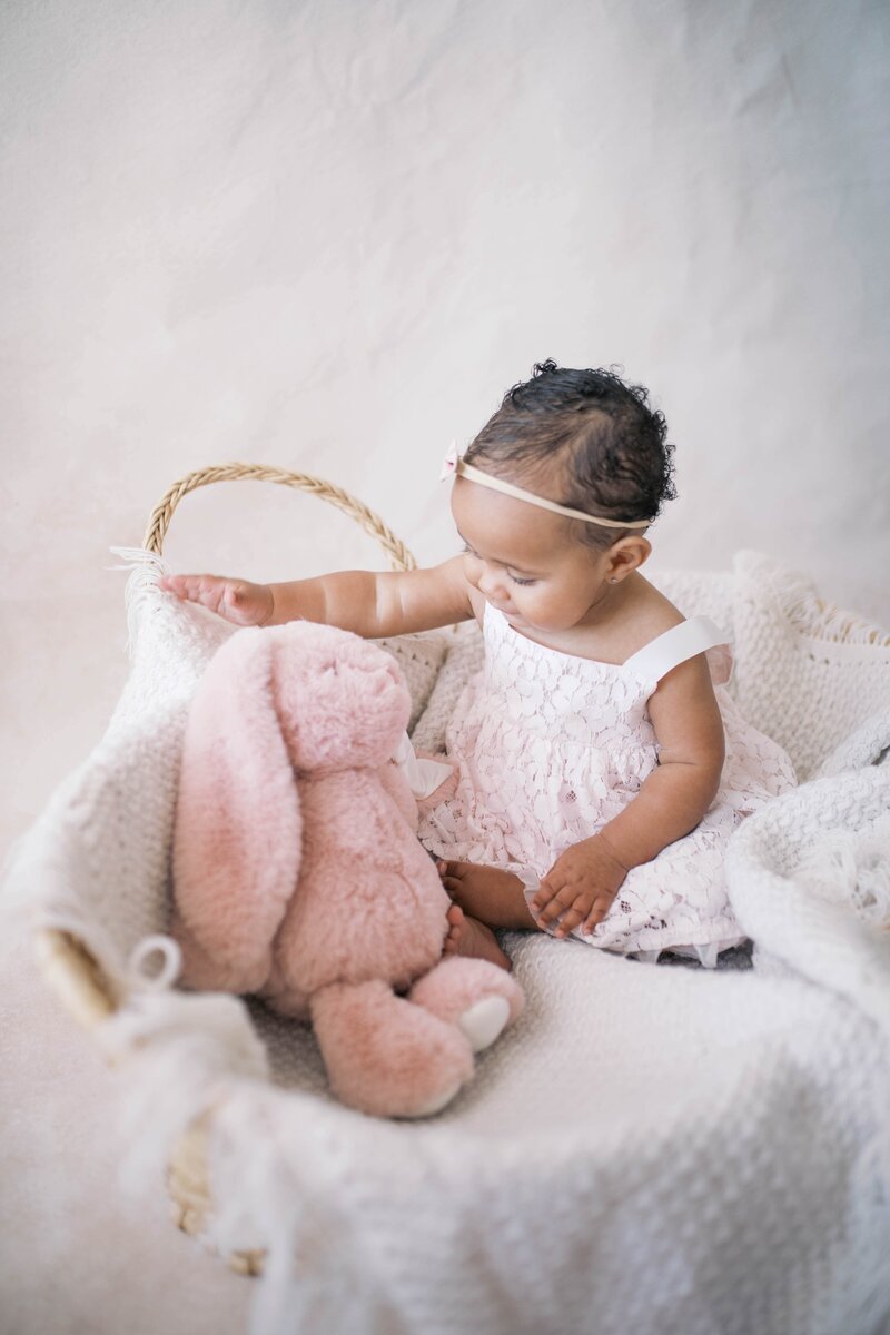 One year old Portrait taken in a studio in Jacksonville with the baby in a blush pink dress playing with a rabbit in her basket