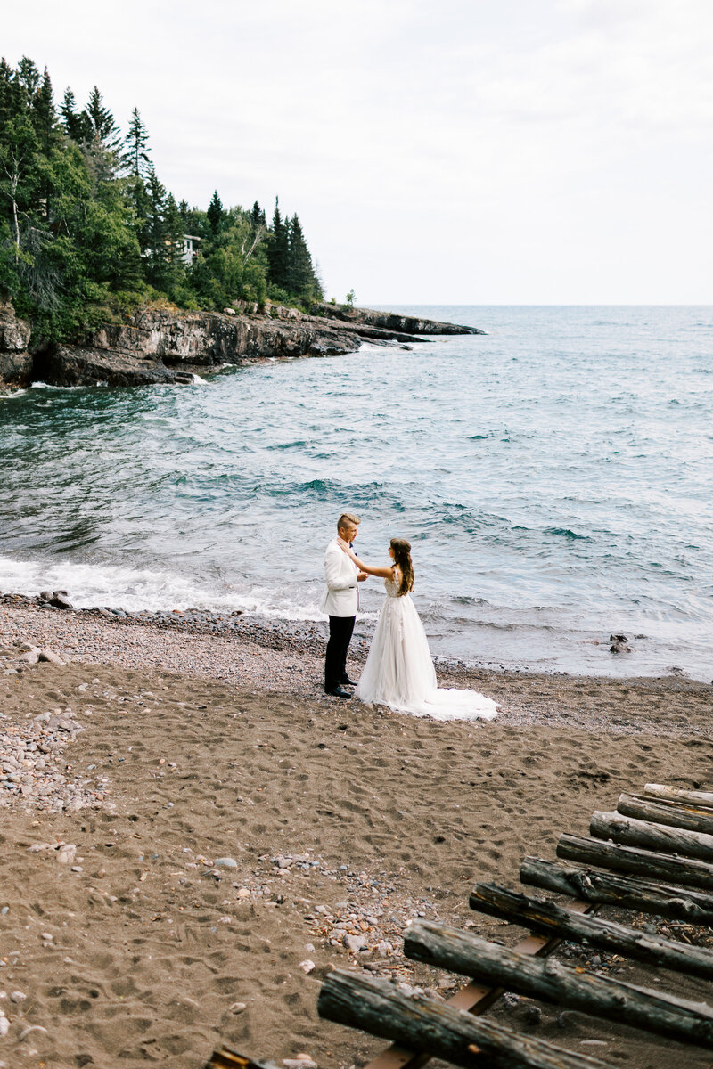 Bride and groom share a romantic moment on the North Shore beach as he reads his vows and she gently wipes his tears, with Lake Superior in the background — captured by Toly Dzyuba Photography, Minneapolis wedding photographer.