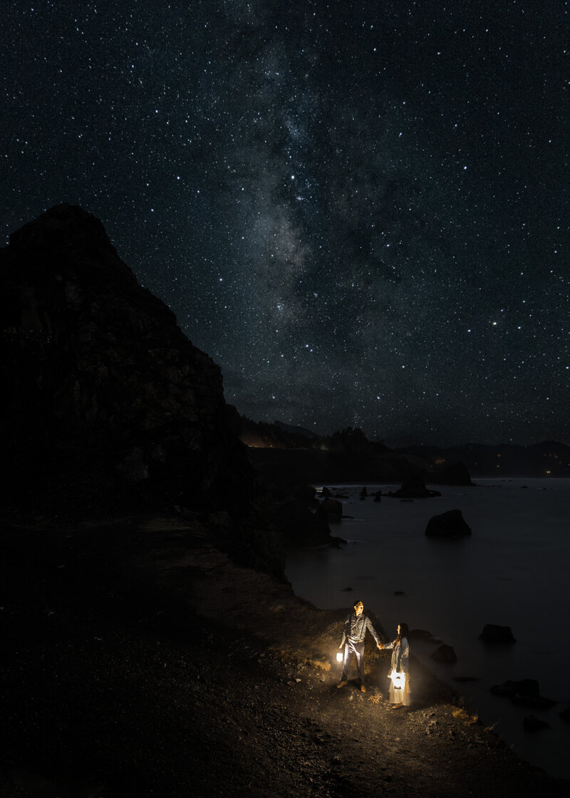 At the end of their Oregon coast elopement, a couple explores the coastline with lanterns. the srs above them are shining .