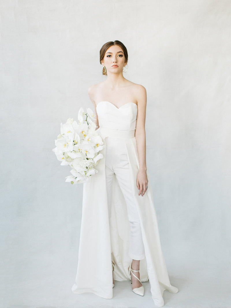 bride wearing pant suit with cape wedding dress holding an all white orchid bouquet