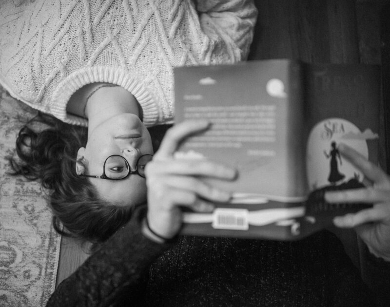 In a black and white photo, a woman with glasses relaxes on the floor, deeply while her partner reads them a book.