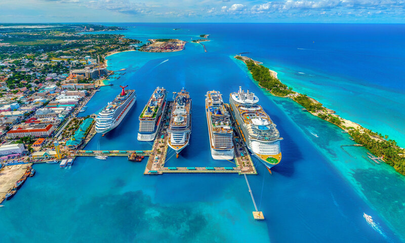 An aerial view of multiple cruise ships lined up in a row at various docks, with the ocean expanse appearing in bright blue and turquoise, and an island to the left side.