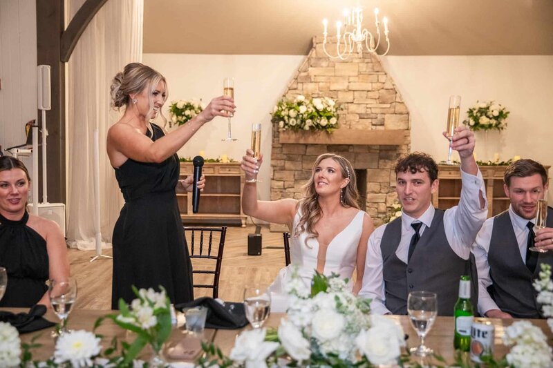 At a reception in Lancaster, a bridesmaid raises a champagne flute during a toast while the newlyweds smile up at her. The bride wears a sleek white dress, and the rustic stone fireplace behind them is adorned with florals. Warm wood tones and intimate lighting create a grounded, celebratory vibe rooted in Central Pennsylvania hospitality.