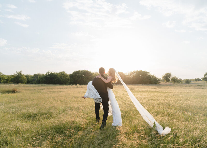 Landscape image of groom carrying bride through grassy field while her veil trails behind them