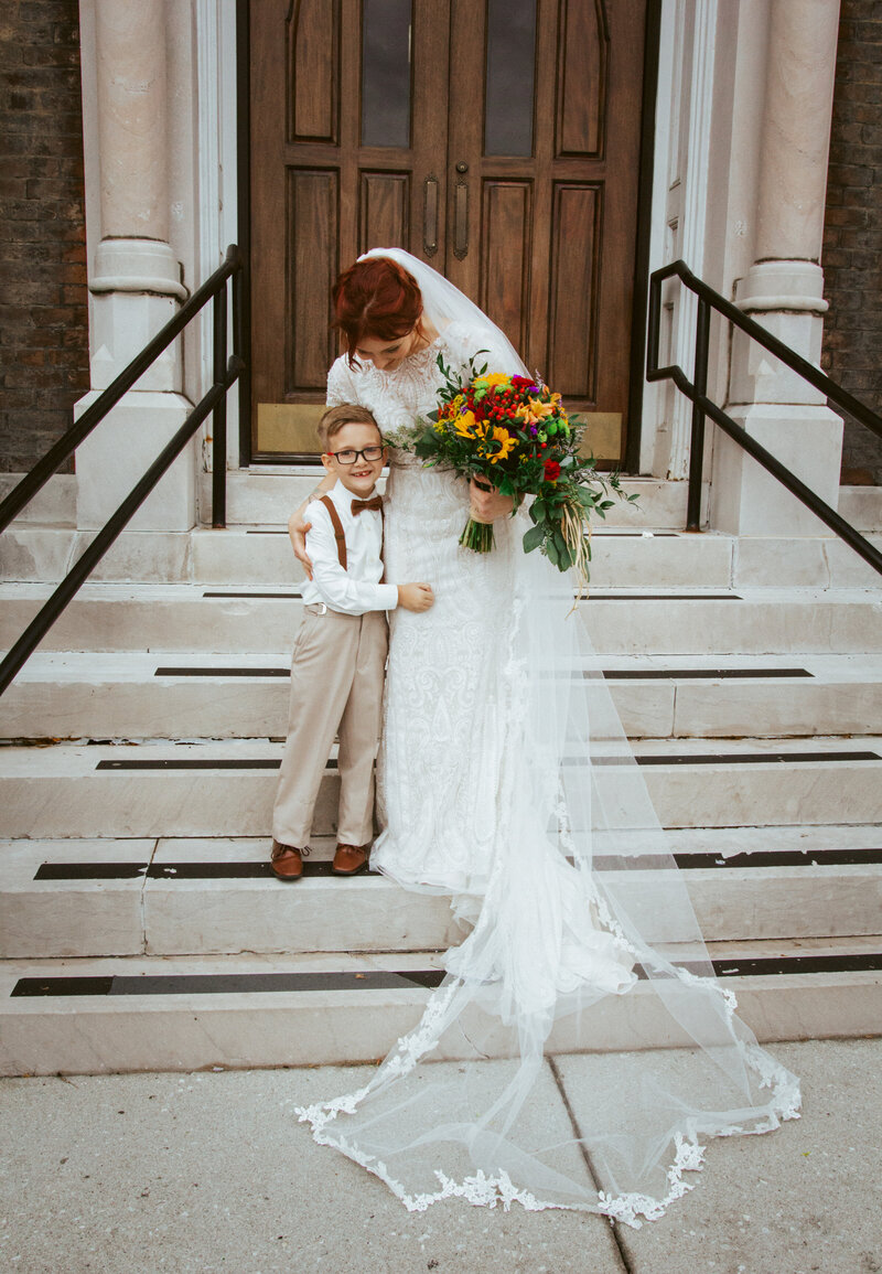 bride in white dress hugging young boy on steps of church