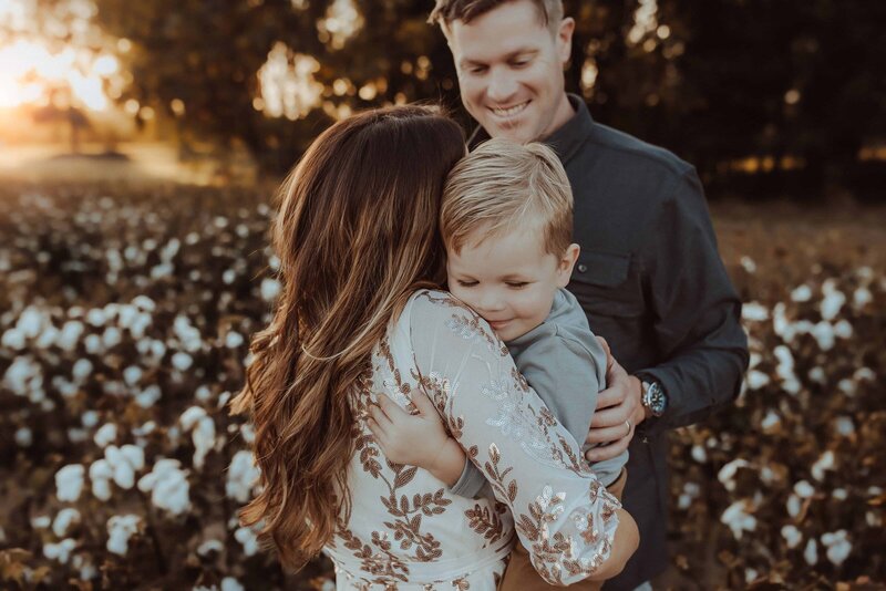 A woman embraces a young boy as a man stands behind them, smiling. Captured by a family photographer in Macon GA, they stand in a field of white flowers with the sun setting beautifully in the background.