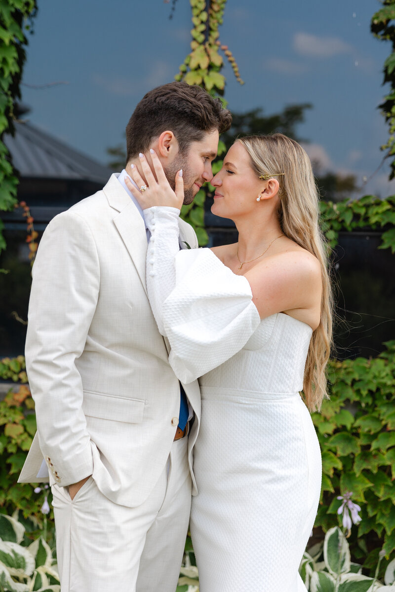 Couple in white attire share intimate moment in garden with greenery backdrop.