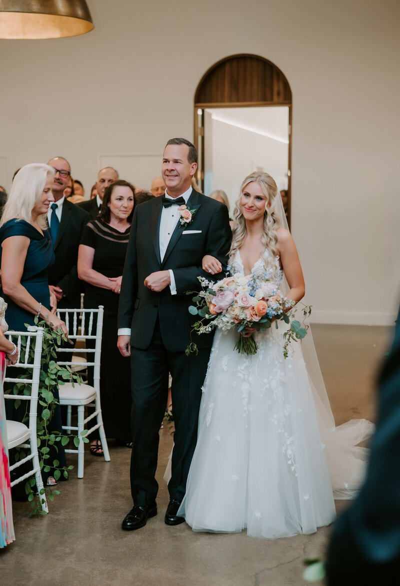 Bride walking down the aisle with her father during an indoor wedding ceremony, holding a bouquet of pastel flowers while guests look on