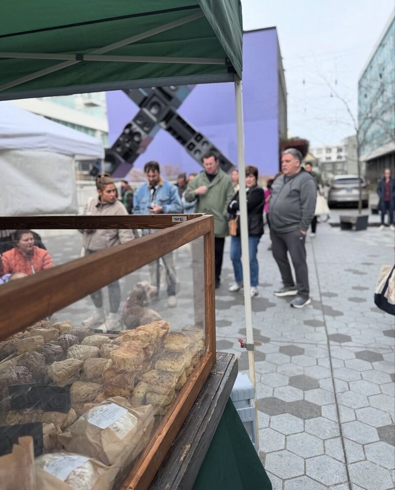 Grain Artisan Bakery’s booth at a Seattle farmers market, featuring rows of gluten-free pastries and breads under a green tent, with customers lining up to shop local and support small-batch baking.