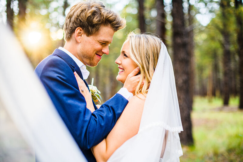Close up of bride and groom smiling looking at each other groom holding bride face cheek golden hour light and pine trees behind them in background, bride's veil in foreground, in Flagstaff Arizona Serendipity Venue