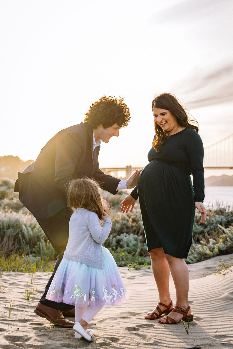 mom standing on beach in front of Golden Gate Bridge as Dad and daughter reach to touch her baby bump