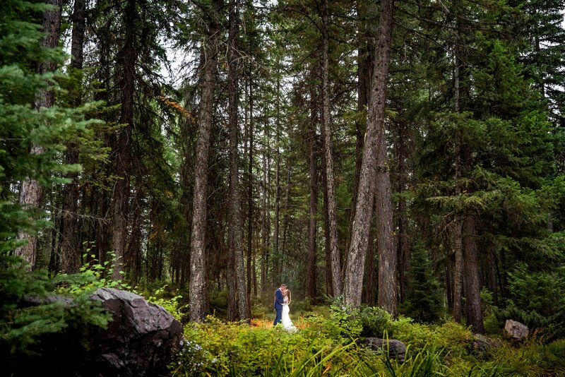 Bride and groom stand close together underneath large grove of pine trees in Montana.