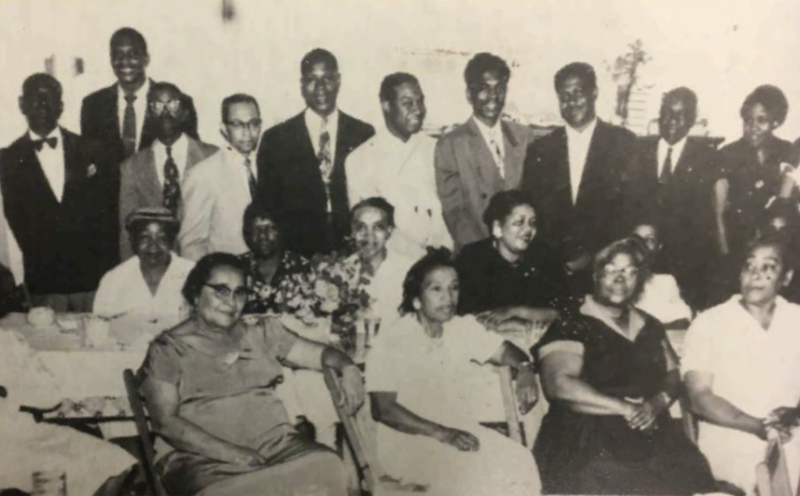 “A vintage black-and-white photo of Black families dressed in formal attire, gathered in a living room—capturing the warmth, resilience, and deep-rooted connection of Black community life in Northwest Arkansas.”