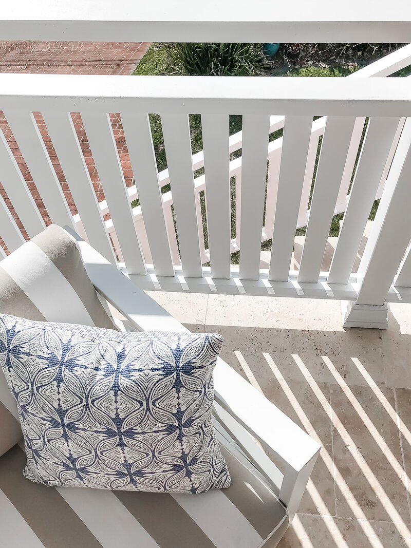 This white chair, adorned with a patterned blue and white pillow, sits gracefully on a sunlit porch beside a white railing. It's an example of bespoke interior design in Newcastle NSW, adding elegance and personalized charm to your outdoor space.