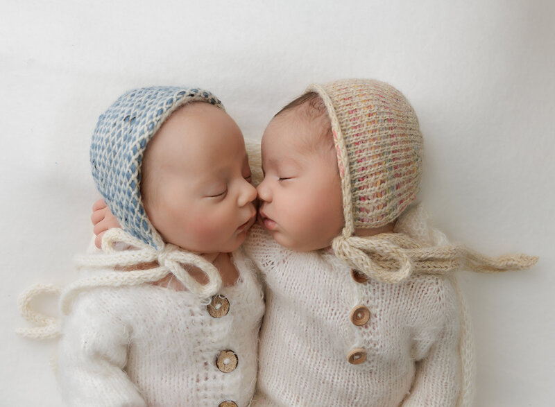 Newborn twin photoshoot  Close-up aerial image of their faces. Baby girl and boy are lying with their noses touching. Babies are sleeping and wearing matching knit rompers and bonnets. Captured by best Brooklyn, NY newborn photographer Chaya Bornstein.