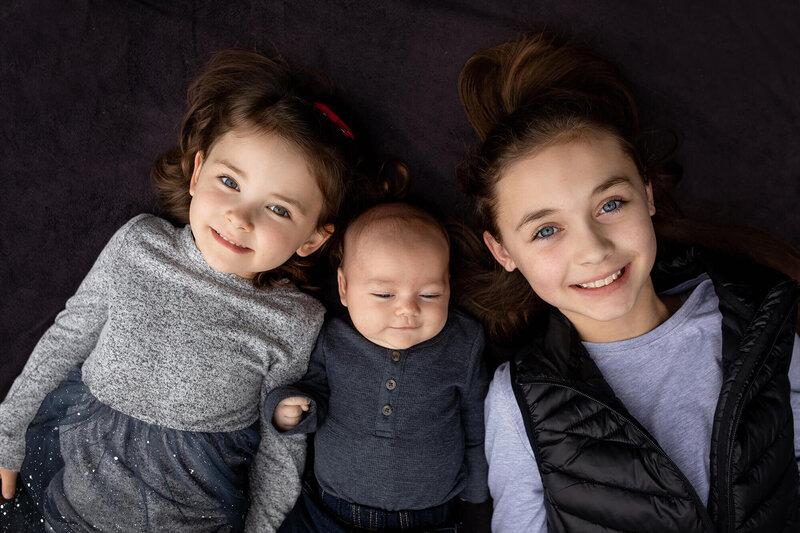 Two sibling girls laying with their little baby brother in between them.