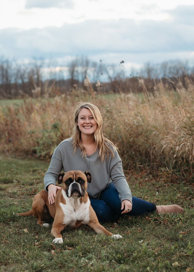 Chiropractor Brittany Fisk sitting in a grassy field with her brown boxer dog, representing Busy Bee Chiropractic’s caring approach.