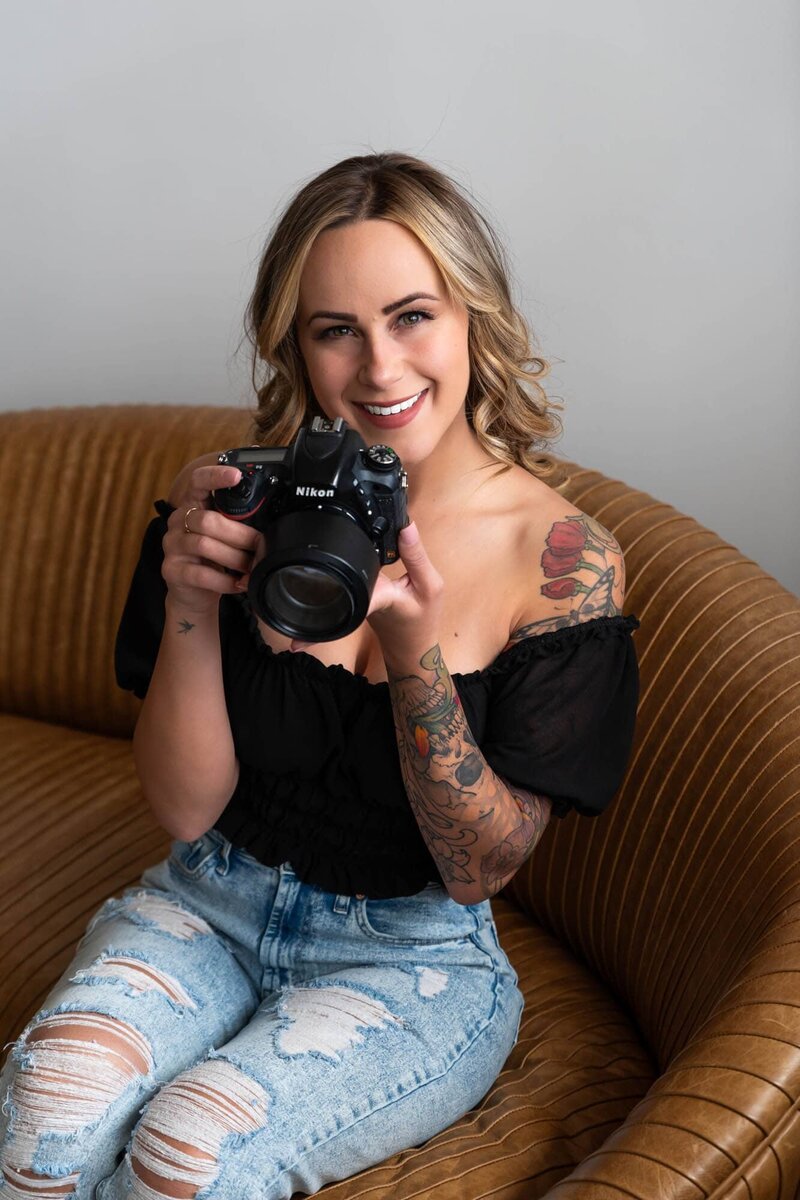 Denver boudoir and wedding photographer smiling with Nikon camera – female photographer with tattoos and blonde hair sitting on a velvet couch, ready to capture intimate, candid moments with confidence and creativity.